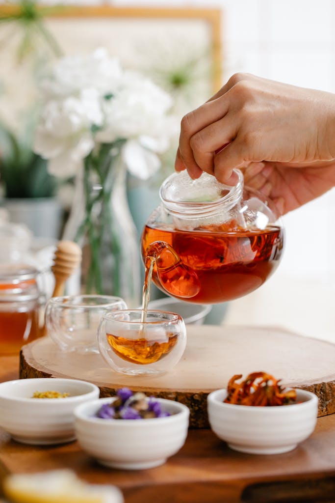Hand pours herbal tea from glass teapot into cup on wooden table with flowers.