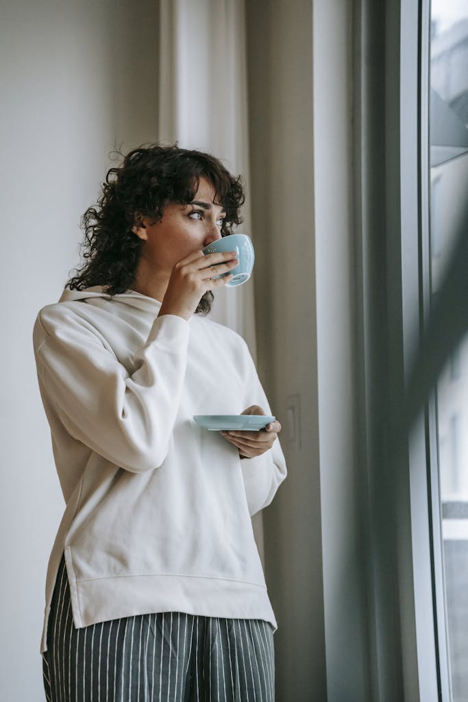 Low angle of female in casual wear drinking hot coffee in morning standing near panoramic window