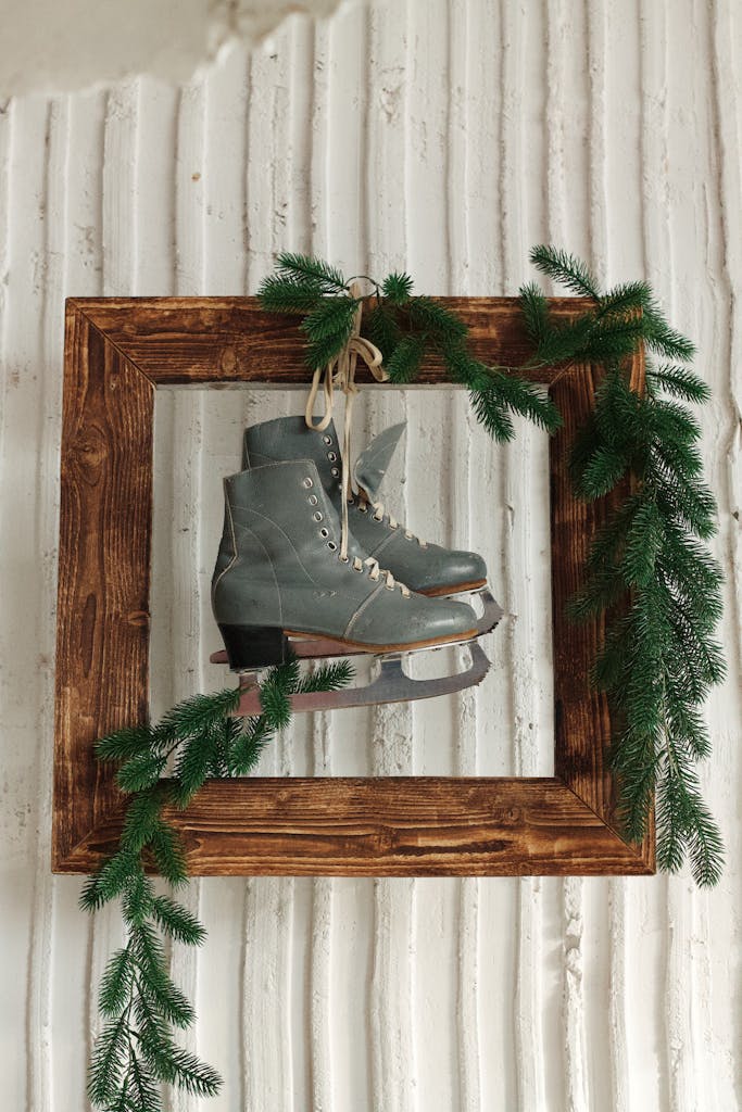Old ice skates in wooden frame with fir branches on rustic white wall.