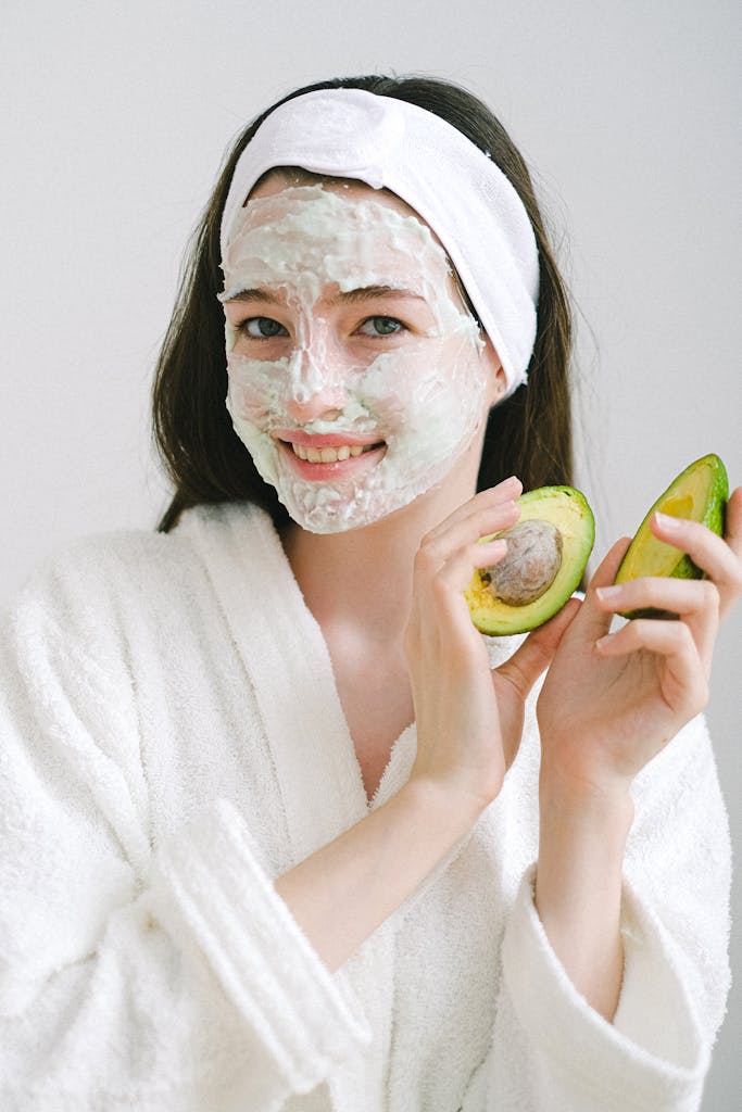 Optimistic female with moisturizing cream on face wearing bathrobe looking at camera while standing on white background with avocado in hands