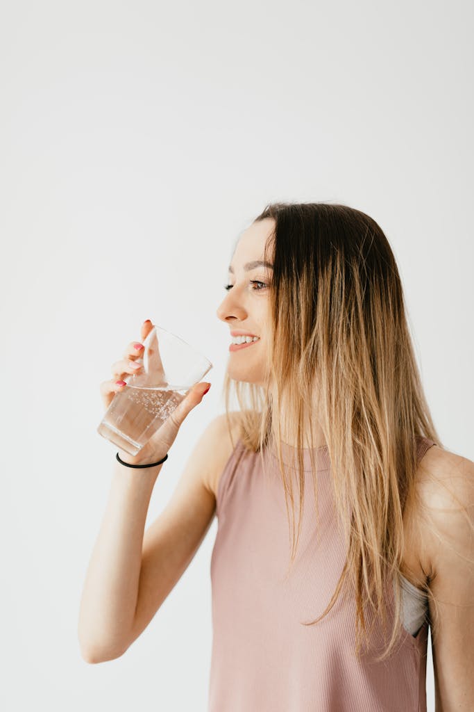 Positive young female in casual wear drinking glass of cold water and looking away against white background