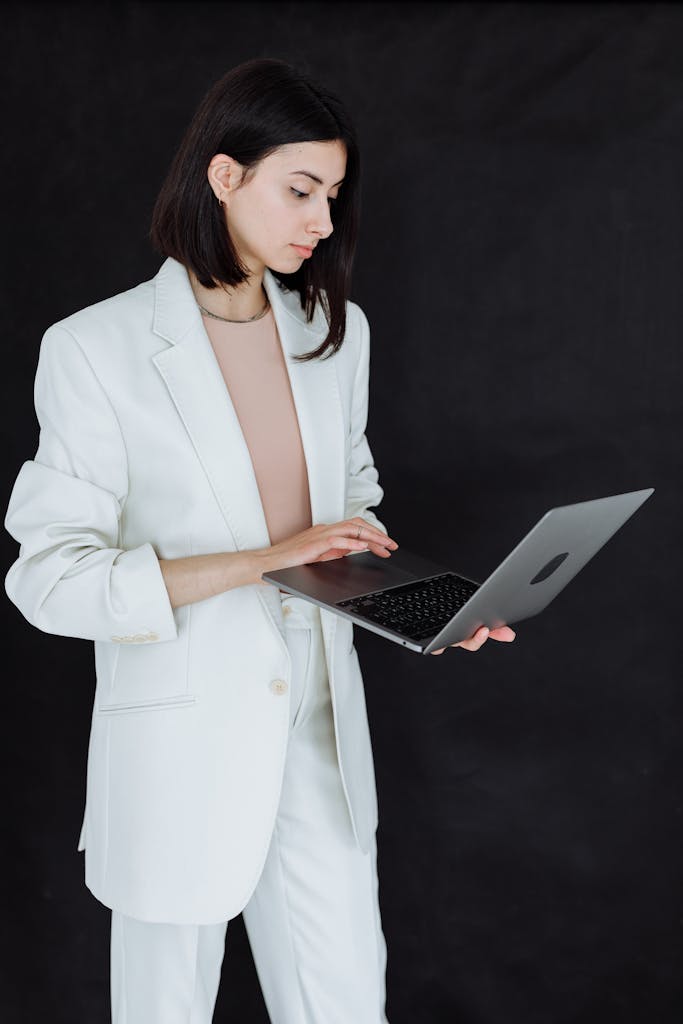 Professional woman in a white blazer using a laptop against a dark background.