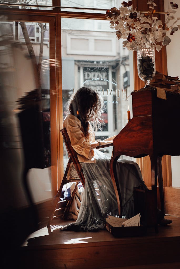 Side view full body of unrecognizable female in retro outfit sitting at wooden writing cabinet in vintage store near window