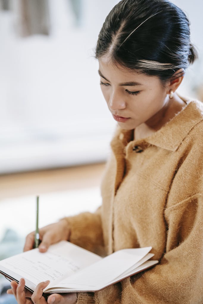 Side view of young Hispanic female in casual clothes with pen reading information from notebook in light room