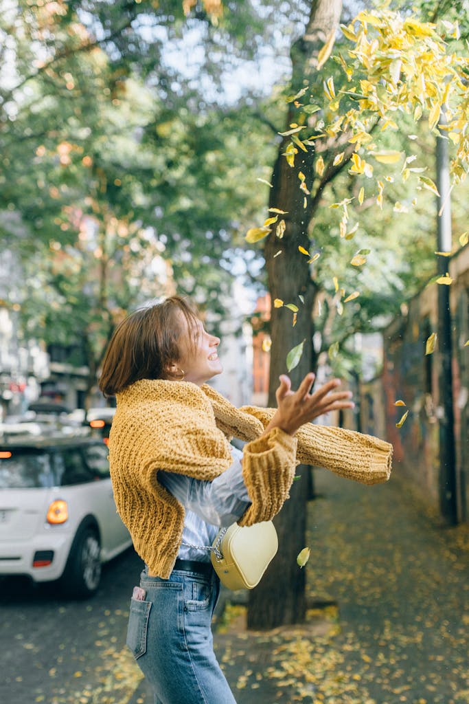 Smiling woman in a sweater tosses autumn leaves on a city sidewalk, capturing fall joy.