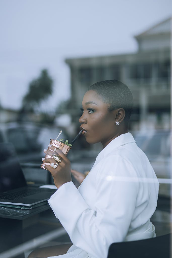 Stylish woman in white sipping a drink while working on a laptop indoors.