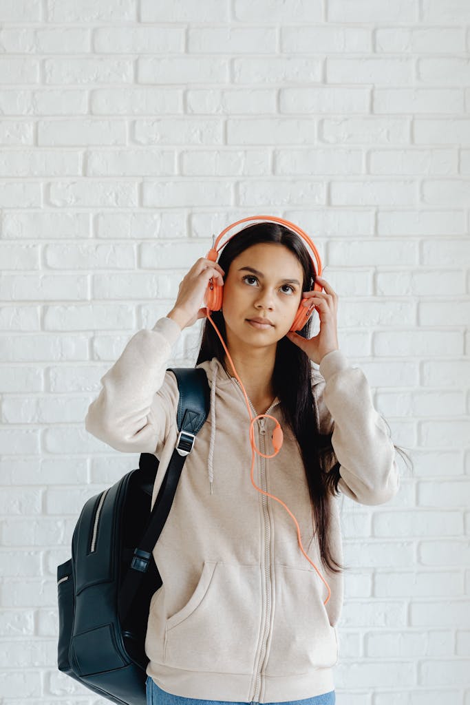 Teenage girl wearing headphones and backpack against white brick wall.