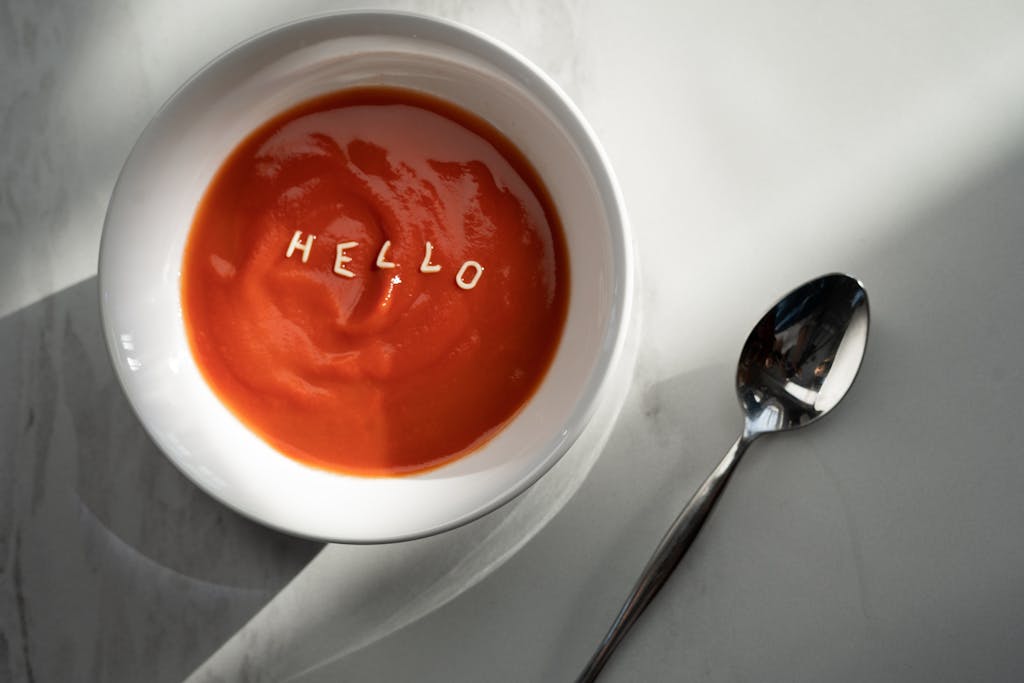Tomato soup in a bowl with the word 'hello' formed by pasta letters, alongside a spoon.