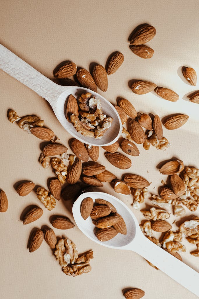 Top view of assorted almonds and walnuts in wooden spoons on a beige background.