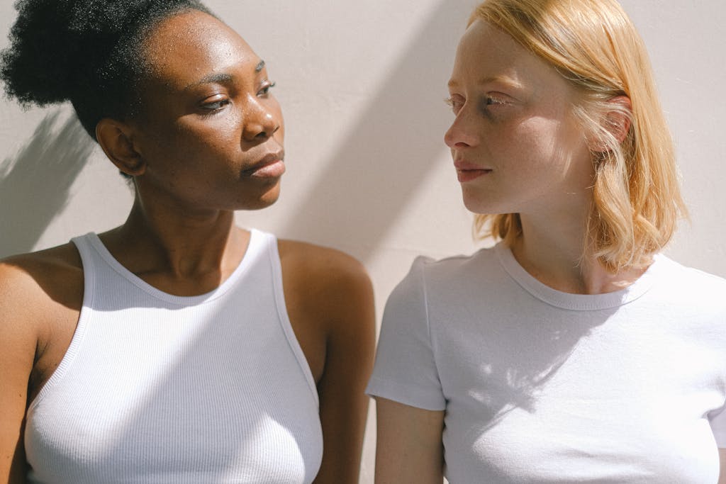 Two diverse women in white tops share a moment of connection in soft daylight.