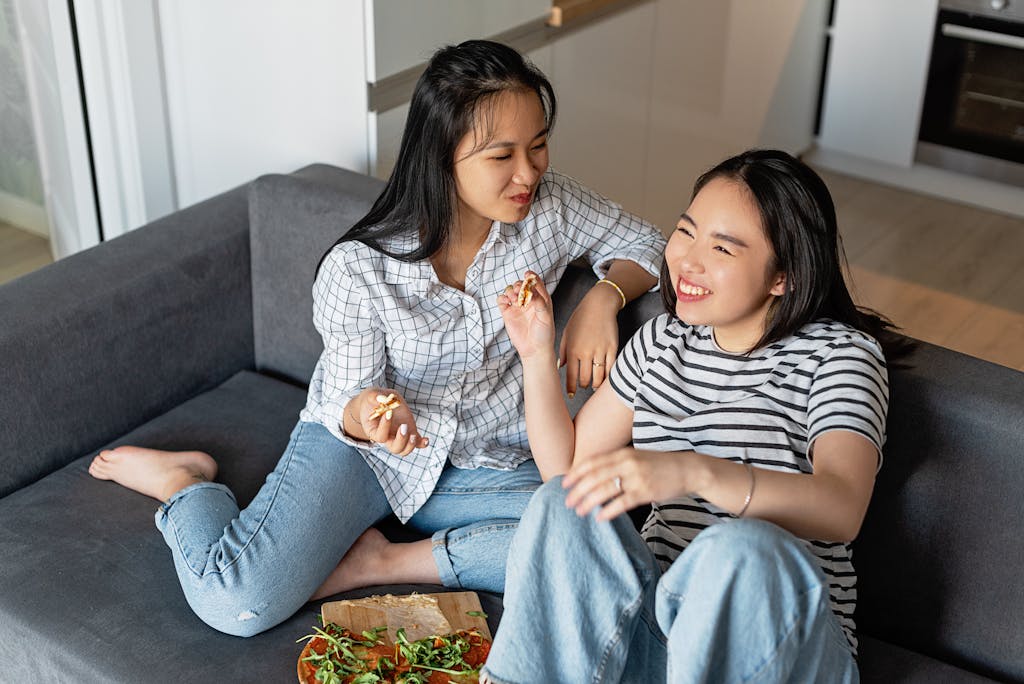 Two friends sit on a sofa sharing pizza, smiling and enjoying a relaxed day indoors.