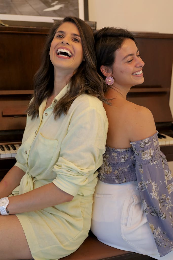Two women laughing and relaxing back-to-back in front of a piano indoors.