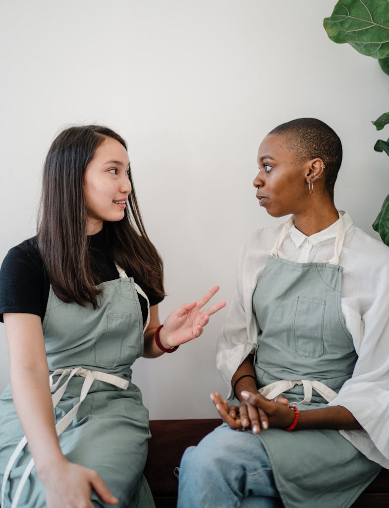 Two women wearing aprons engaged in a casual conversation indoors, conveying teamwork.