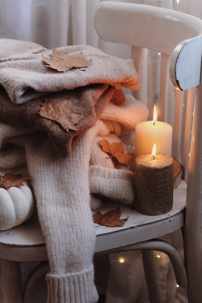 Warm autumn vibes with lit candles on a chair surrounded by cozy sweaters and dry leaves.