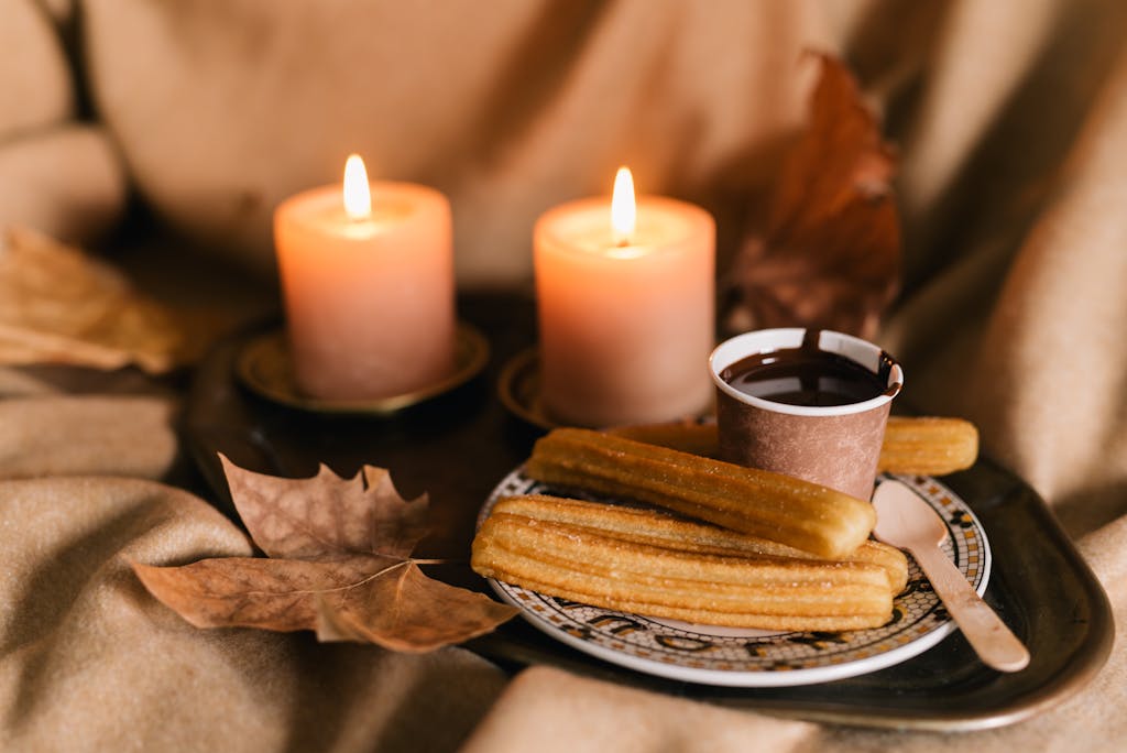 Warm churros and chocolate surrounded by lit candles and autumn leaves, creating a cozy fall atmosphere.