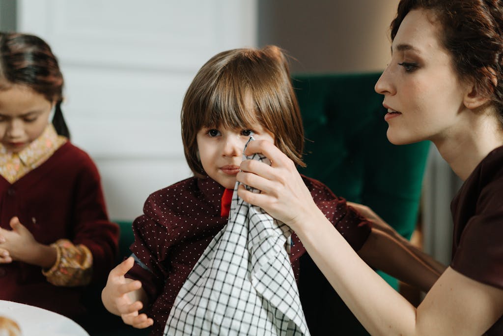 Woman assisting a child with a napkin during a meal indoors, highlighting care and family bonding.