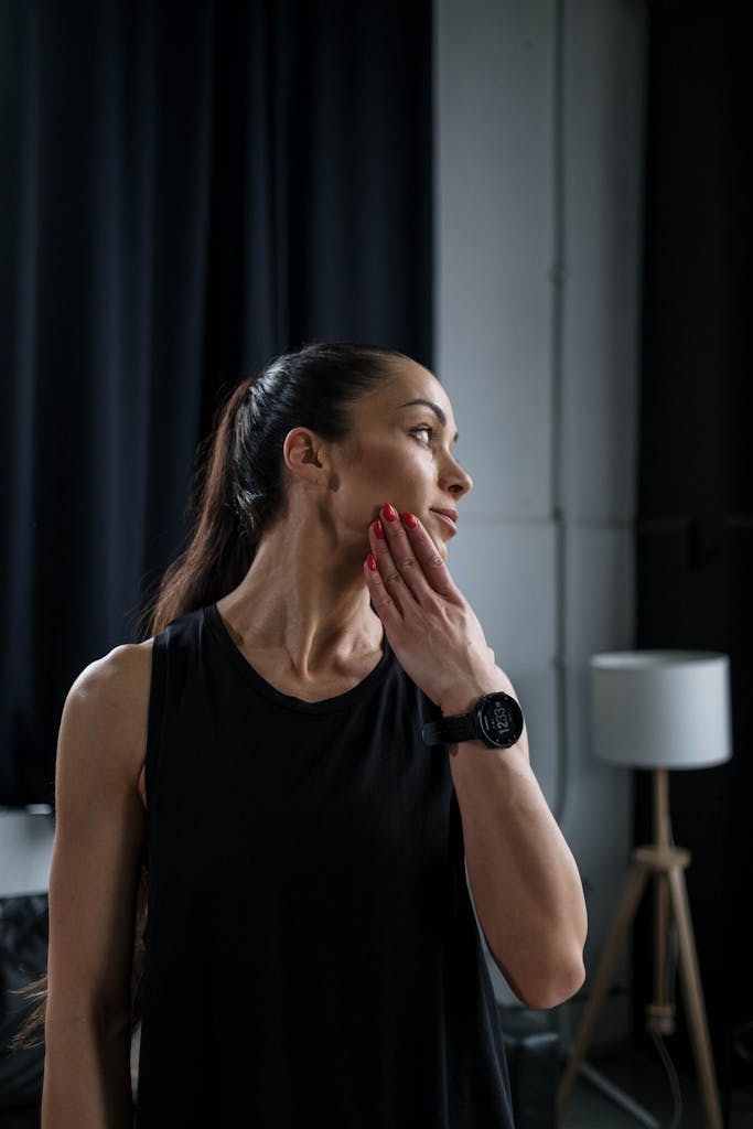 Woman in black workout attire stretching indoors with hand on chin.