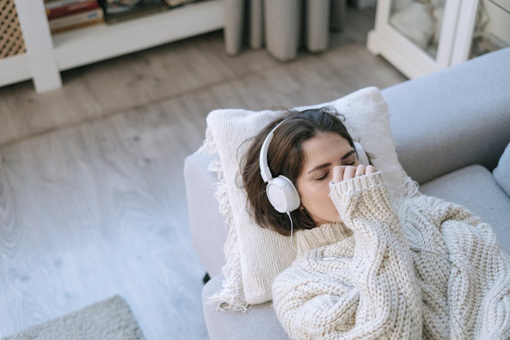 Woman in cozy sweater with headphones relaxing on sofa, eyes closed, enjoying music.