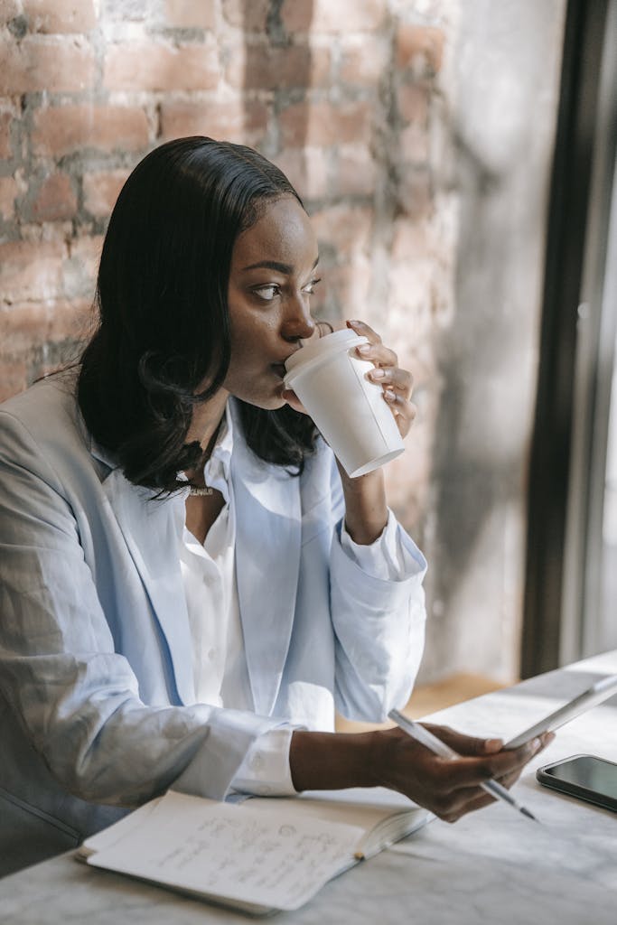Woman in professional attire drinking coffee, sitting by a brick wall with a tablet and notebook.
