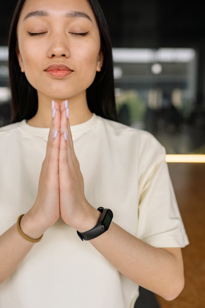 Woman in white shirt meditating indoors with hands together, eyes closed.