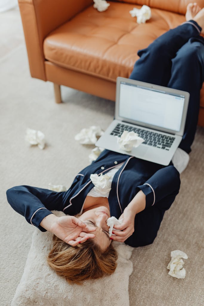 Woman lying on the floor with a laptop, surrounded by used tissues, looking stressed or sad.