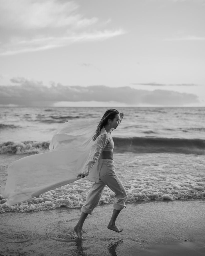 Woman walking by the sea in a serene black and white beach scene at sunset.