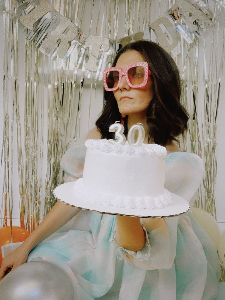 Woman wearing pink glasses holding a 30th birthday cake at a party.