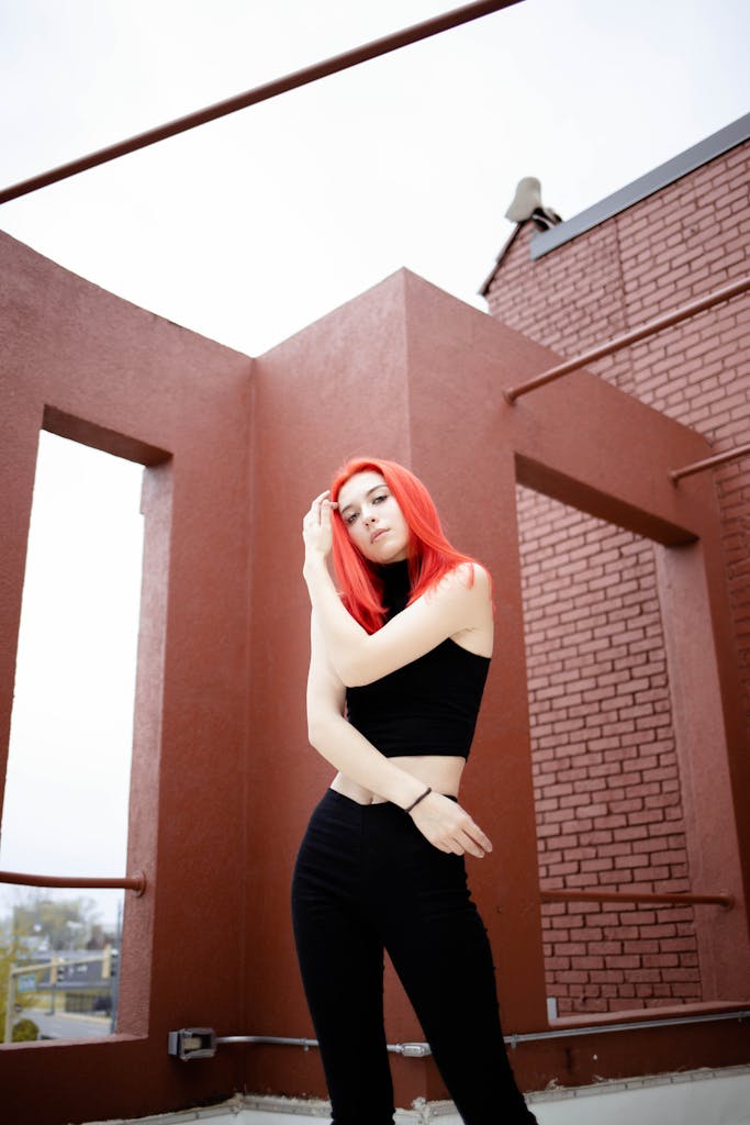 Woman with red hair posing in stylish black outfit against brick structure.