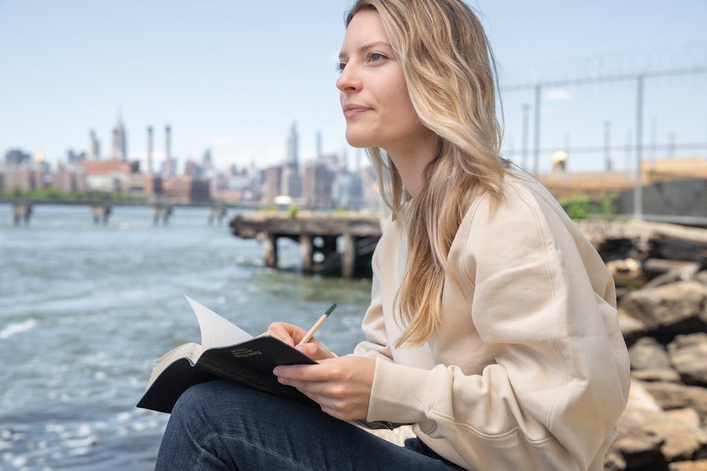 Woman writes in a notebook by the water with a city skyline in the background.