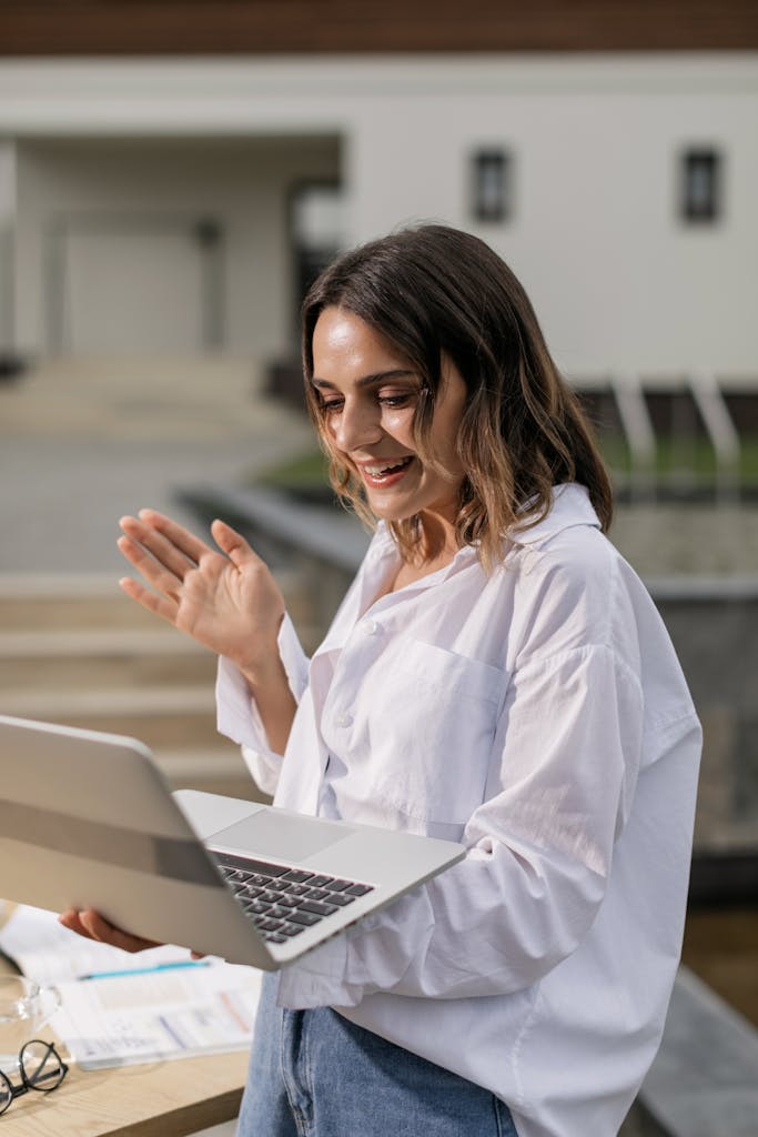Young woman in white shirt smiling during a video call on her laptop outside.