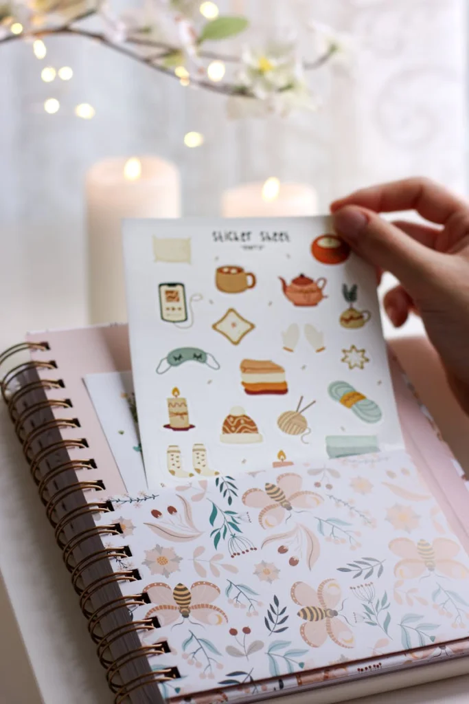 A hand holding cute stickers above a floral notebook with warm candlelight.