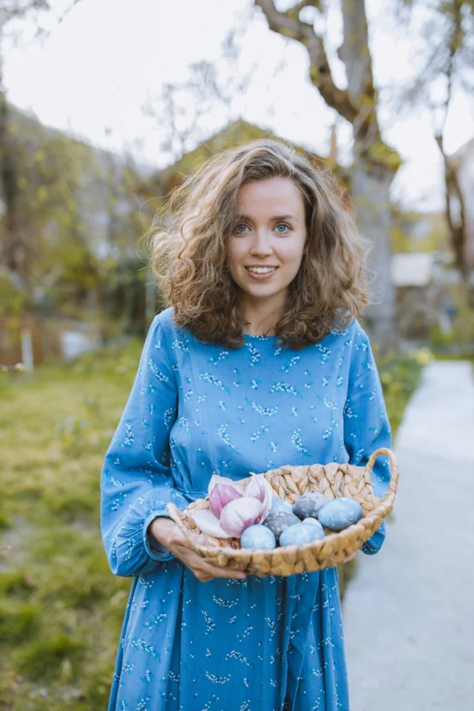 Smiling woman holding Easter basket with painted eggs in a sunny park setting.