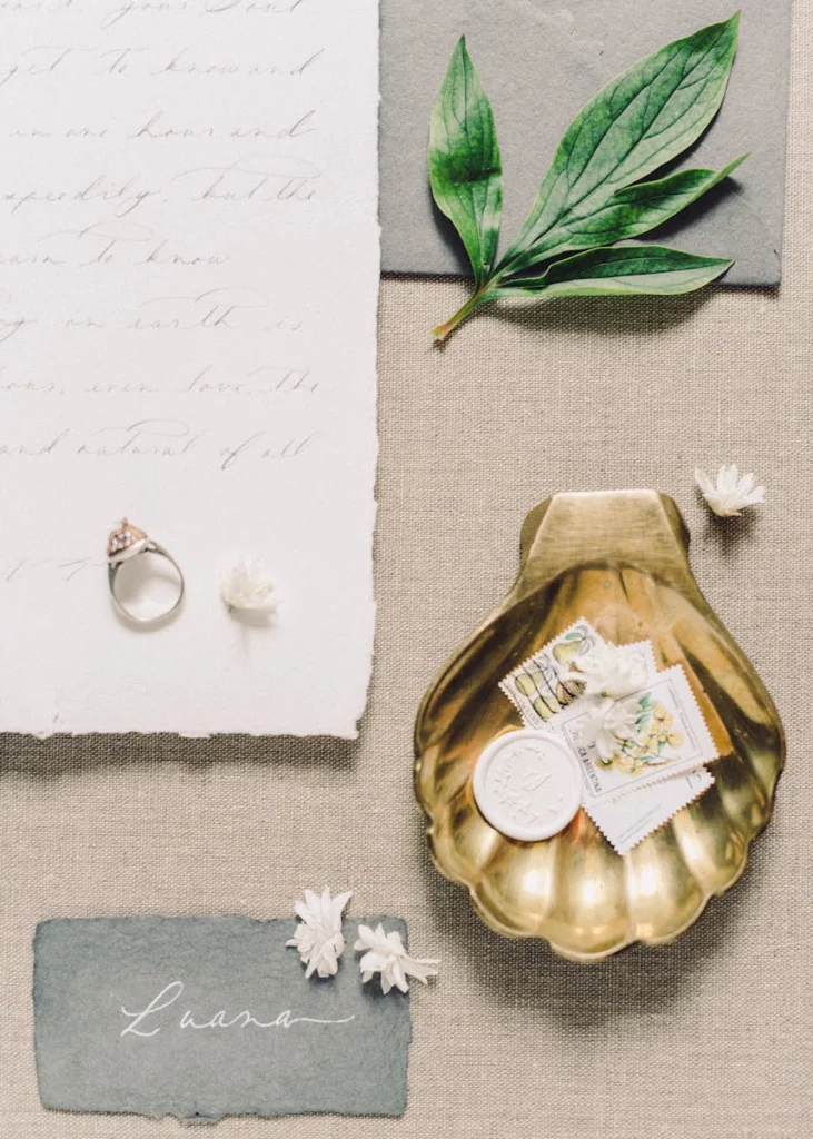 Elegant still life featuring a golden shell tray with stationery, leaves, and a ring on a textured surface.