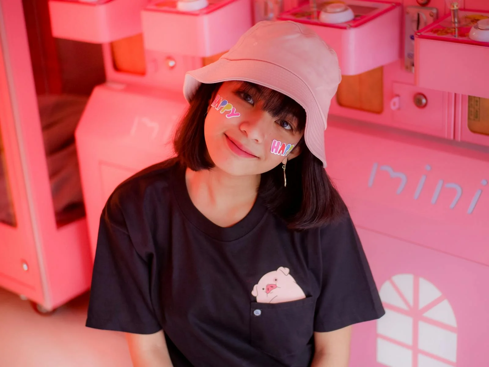 Smiling young woman with 'happy' stickers on her cheeks indoors wearing a hat.