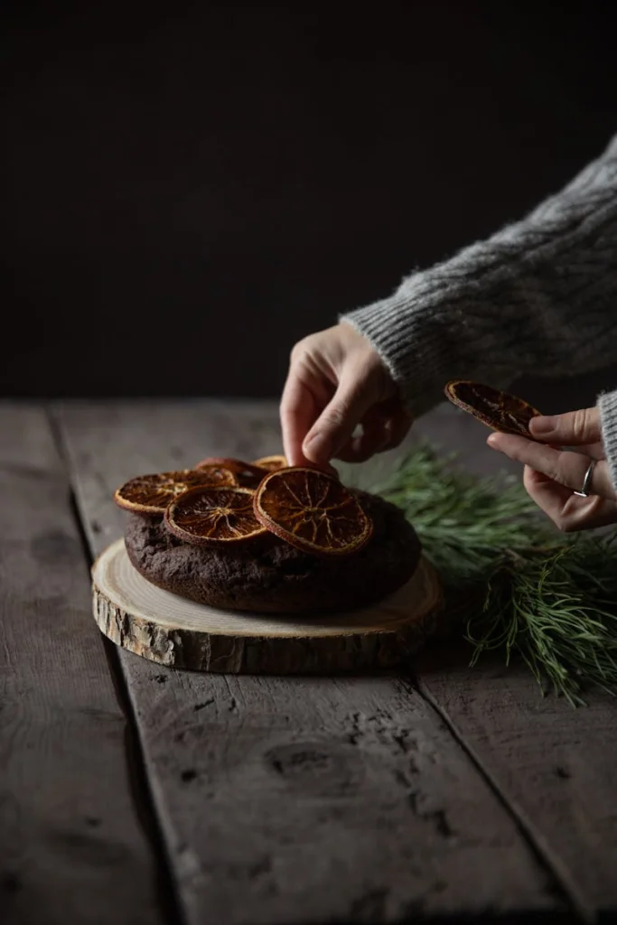 Hands decorating chocolate cake with dried oranges on wood slice.