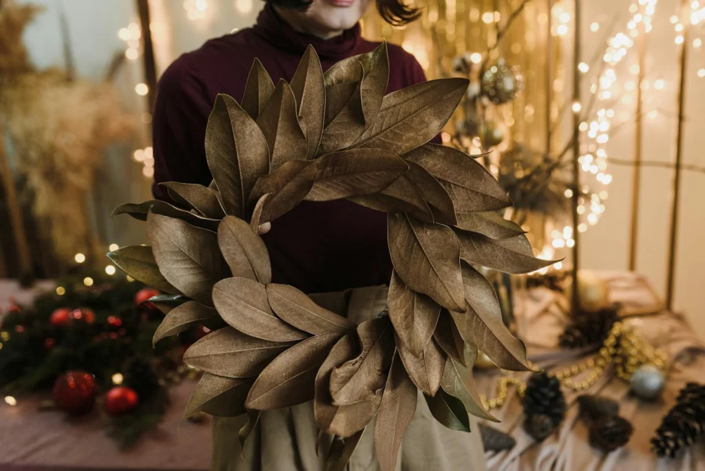 Woman holding a handmade wreath surrounded by festive holiday decorations indoors.
