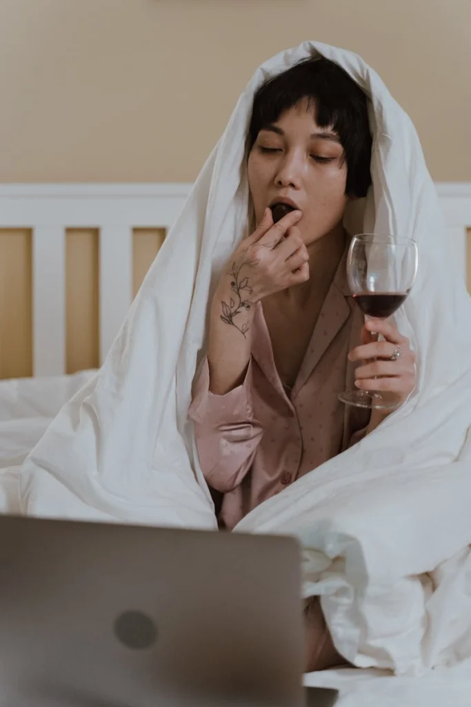 Asian woman in pajamas enjoying wine and chocolate while sitting in bed, wrapped in a blanket.