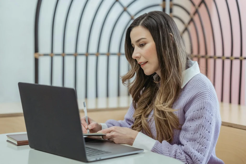 Focused female freelancer with wavy hair writing notes in planner while sitting at table and working on netbook
