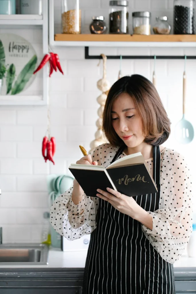 Elegant Asian woman in apron taking notes in a modern kitchen setting.