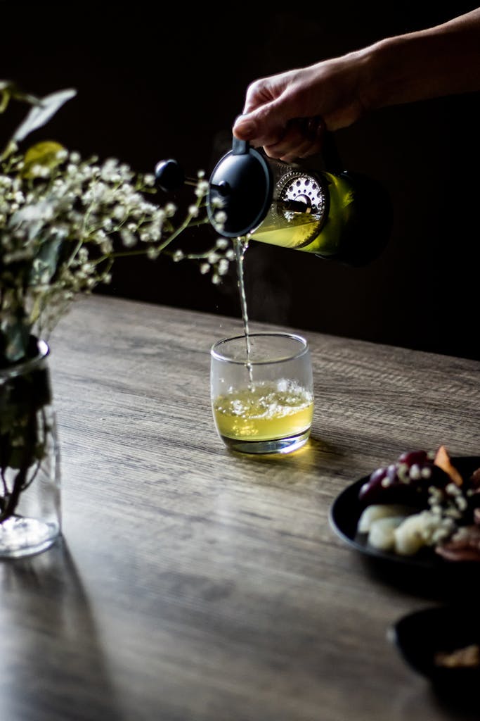 A close-up shot of herbal green tea being poured into a drinking glass on a wooden table, with flowers and snacks.