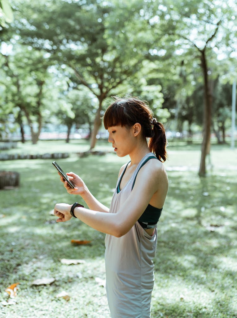 A fit young woman checks her smartwatch while holding a smartphone in a serene park setting.