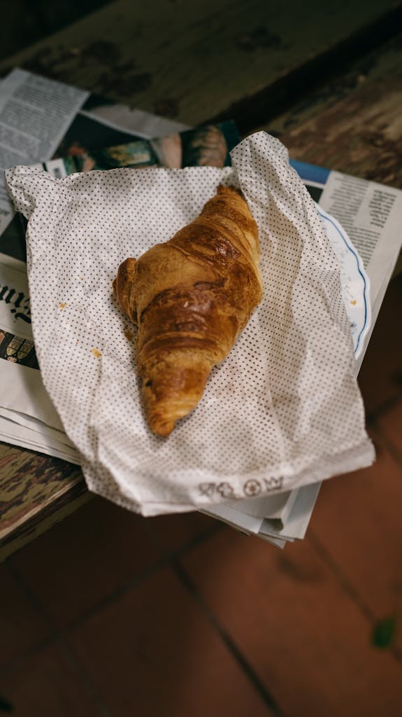 A golden-brown croissant on paper with newspaper background, offering a rustic breakfast vibe.