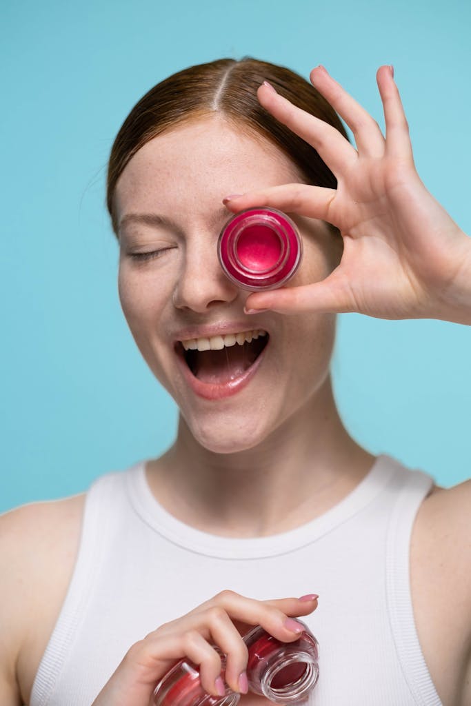 A joyful woman holds lip balm to her eye against a vibrant blue background in a studio setting.