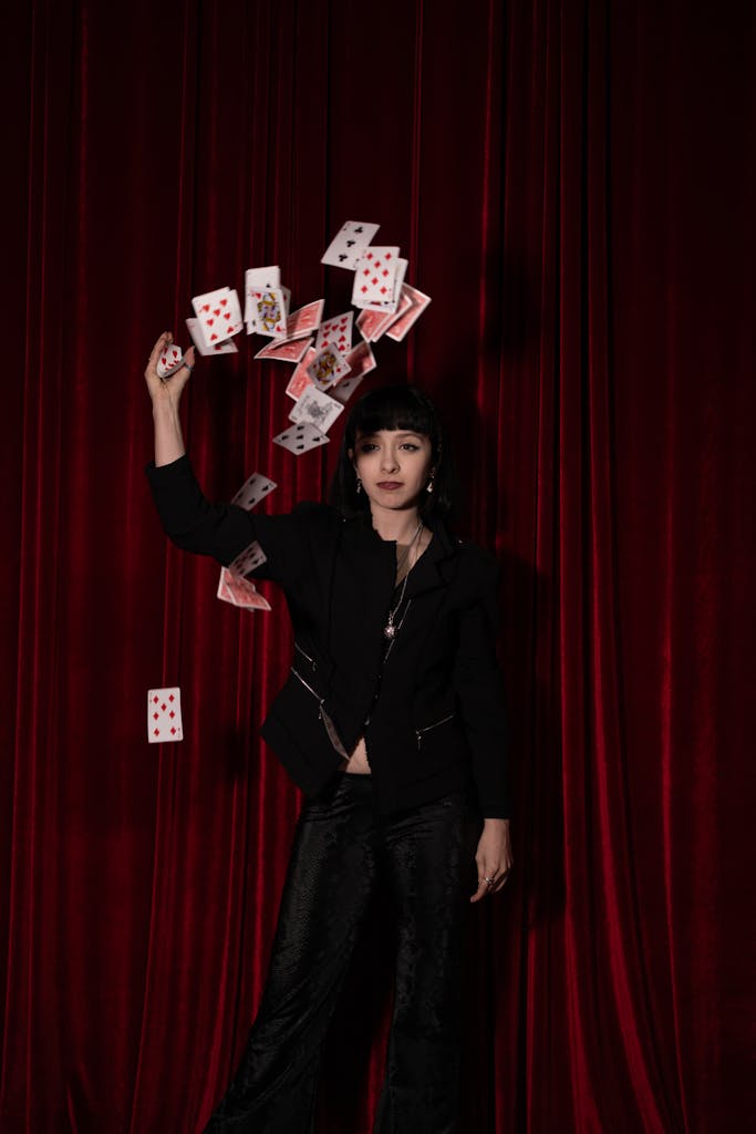 A magician in a stylish outfit throws playing cards against a red curtain backdrop.