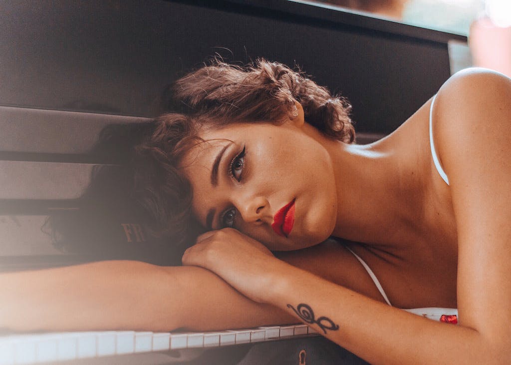 A thoughtful woman in a relaxed pose with striking red lipstick rests her head on a piano.