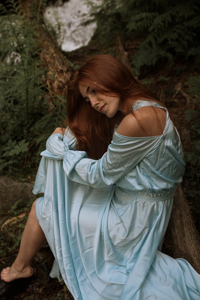 A woman in a flowing blue dress sitting peacefully amidst lush green forest foliage.