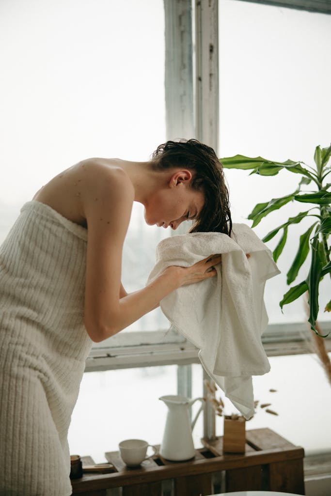 A woman in a towel dries her hair by a window, creating a serene and fresh morning routine scene.