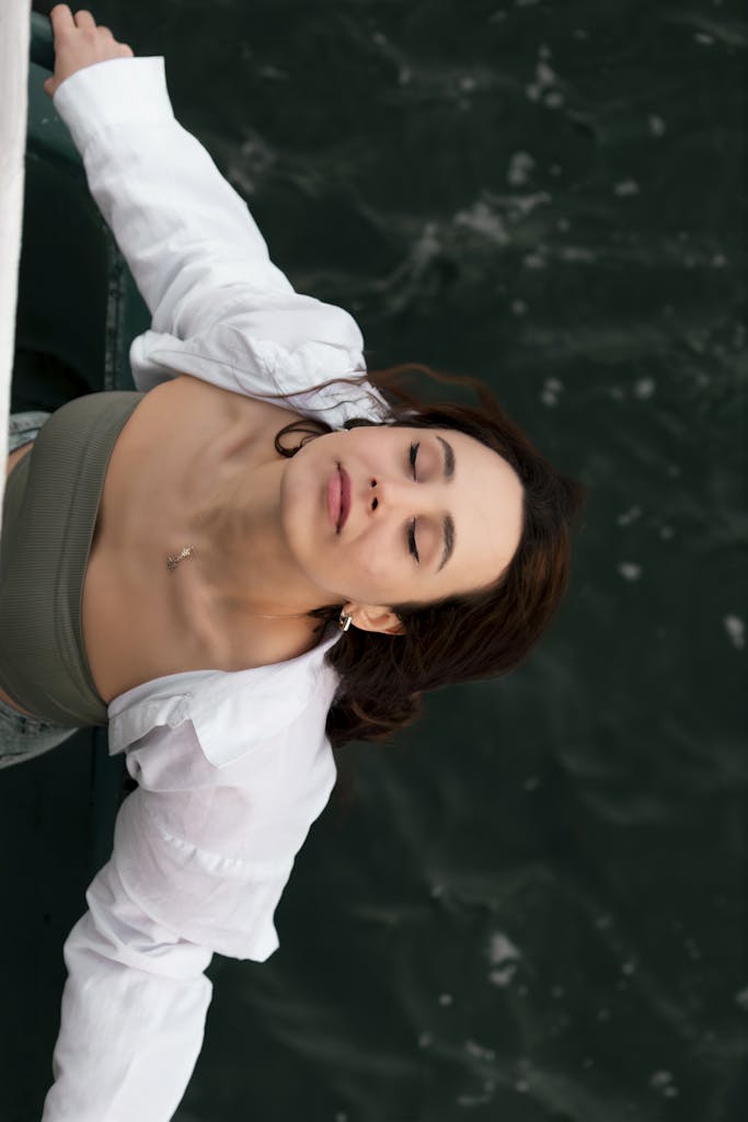 A woman in a white top enjoys a serene moment outdoors with her eyes closed.