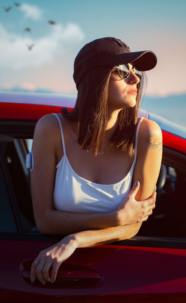 A woman in sunglasses leans out of a red car window, enjoying the sunny outdoors with mountains in the background.