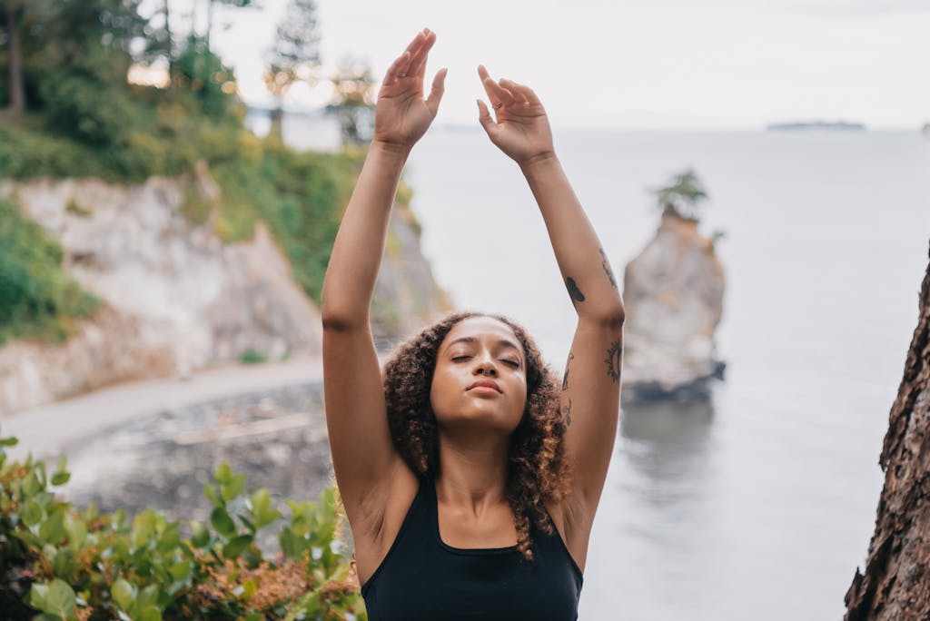 A woman meditates with arms raised in a serene outdoor cliffside setting, eyes closed in relaxation.